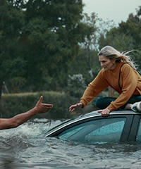Woman on the hood of a car surrounded by water. A hand is reaching out to help her.
